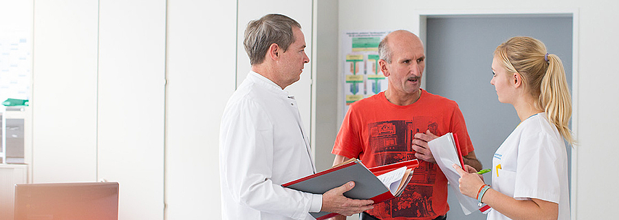 Der Chefarzt der Rehaklinik Klausenbach und eine Pflegefachfrau unterhalten sich mit einem Patienten.