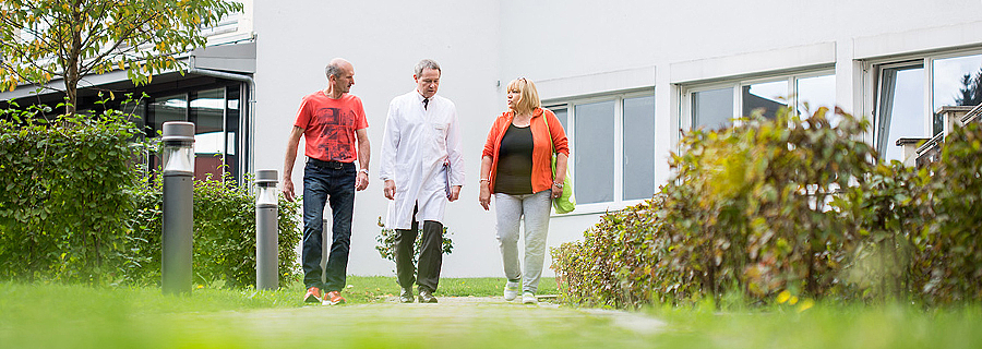 Eine Patientin und ein Patient spazieren gemeinsam mit dem Chefarzt der Rehaklinik Klausenbach im Klinikpark und unterhalten sich mit ihm.