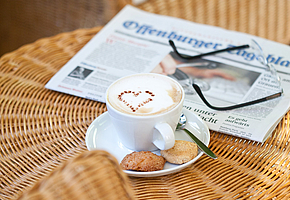 Eine Tasse Kaffee steht auf einem kleinen Tisch, daneben liegt eine Brille auf einer Zeitung.
